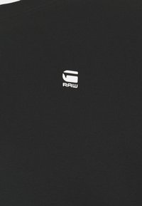 Un tissu noir présente un petit logo blanc "G RAW" centré sur la partie supérieure, suggérant une marque de vêtements décontractés.