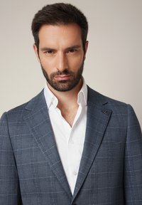 Granatowa marynarka w kratę z szpiczastym klapem, teksturowana tkanina i mały akcent na klapie, zestawiona z białą koszulą pod spodem.