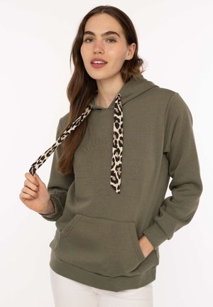 Junge Frau trägt einen olivgrünen Hoodie mit Leopardenmuster-Kordeln, eine Hand hält eine Kordel, die andere in der Fronttasche.