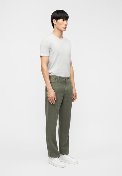 Νέος άνδρας σε πλάγια στάση φορώντας ανοιχτό γκρι κοντομάνικη μπλούζα με V λαιμόκοψη, χακί παντελόνι και λευκά αθλητικά παπούτσια μπροστά σε απλό φόντο.