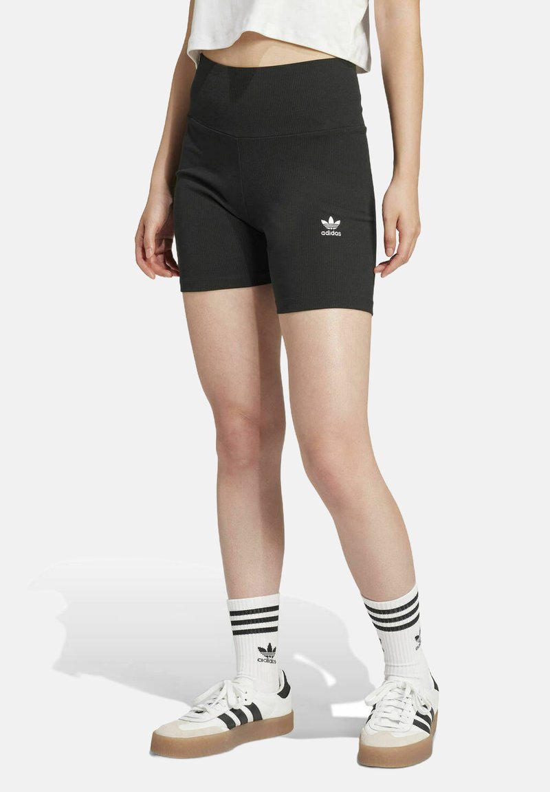 Fekete, bordás rövidnadrág magas derékrésszel és fehér adidas logóval. Fehér és fekete sportcipővel, valamint csíkos térdzoknival viselve.