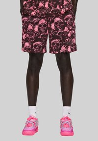 Mand i pink og sorte shorts med dødningehovedmønster, hvide sokker og pink atletiske sko foran en simpel lysegrå baggrund.