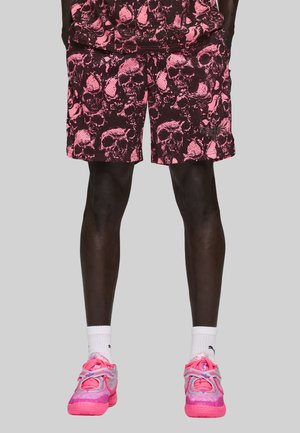Mand i pink og sorte shorts med dødningehovedmønster, hvide sokker og pink atletiske sko foran en simpel lysegrå baggrund.