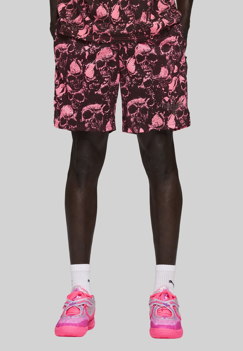Mand i pink og sorte shorts med dødningehovedmønster, hvide sokker og pink atletiske sko foran en simpel lysegrå baggrund.