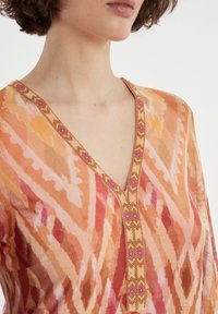 Leichte, gemusterte Bluse in verschiedenen Orangetönen und Pink mit tiefem V-Ausschnitt und dekorativem Besatz mit Perlen entlang des Ausschnitts.