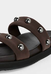 Καφέ σανδάλι με δύο φαρδιές λουρίδες διακοσμημένες με στρογγυλές ασημένιες τρουκς και μαύρη μαξιλαρωμένη σόλα, εμφανιζόμενο σε κοντινή λήψη.