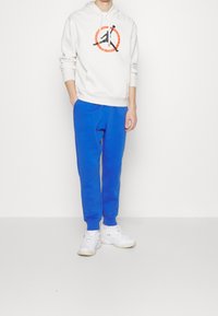 Vit hoodie med en grafisk logotyp, kombinerad med blå sweatpants. Vita sneakers fulländar outfiten. En enkel bakgrund.