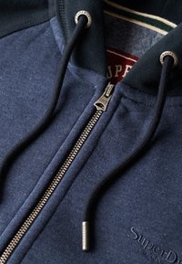 Sudadera con cremallera color azul marino, con una textura suave, cremallera de metal plateado y cordones negros. Presenta bordado del logo en el pecho.