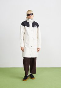 Manteau long imperméable blanc avec section supérieure noire, capuche attachée, poches avant et logo. Porté sur un pantalon en velours côtelé marron foncé et des chaussures noires.