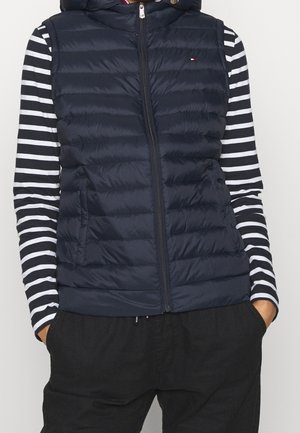 Personne portant un gilet matelassé bleu marine sur une chemise à manches longues rayée noire et blanche et un pantalon noir, les mains dans les poches.