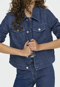 Giacca di denim blu scuro con due tasche frontali, bottoni in metallo argentato e colletto. Le maniche sono arrotolate, mettendo in mostra la consistenza del tessuto.