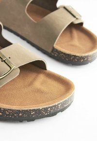 Sandalias de ante marrón que presentan dos correas anchas, una hebilla de metal decorativa y una suela texturizada negra con manchas para mayor agarre.