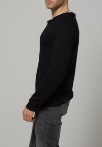 Pull en tricot noir à manches longues avec un col effiloché, un design ajusté et des poignets côtelés, associé à un jean gris.