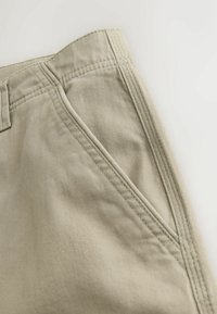 Nærbillede af beige stofbukser, der viser en skrå sidelomme og detaljeret syning langs taljen og lommeåbningen.