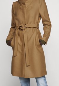 Manteau ceinturé de couleur camel en tissu texturé, doté d'un col haut, de poches latérales et de poignets verts contrastants.
