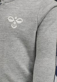 Grå stickad tröja med en subtil broderad bi-logotyp i vitt. Mjuk textur med ribbade muddar. Inga andra dekorationer eller mönster.