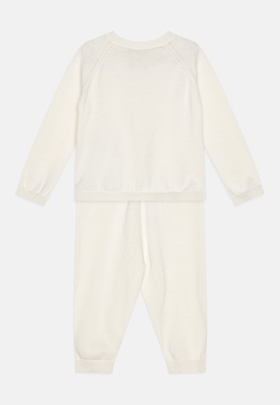 Krēmkrāsas adīta divdaļīga komplekta komplekts, kurā ir garām piedurknēm tops un saskaņotas bikses, abiem ir ribotas aproces un jostasvieta. Mīksta tekstūra.