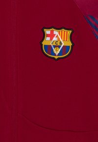 Bordeauxfarvet jakke med FC Barcelona-logo og rød, gul og blå striber; glat tekstur og lynlås lukning.