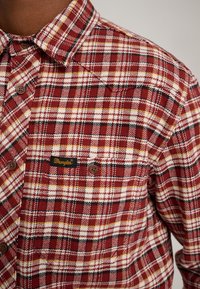 Camicia di flanella a quadri rossi e bianchi con accenti gialli e neri, caratterizzata da un design con bottoni e una toppa con il logo Wrangler sul petto.