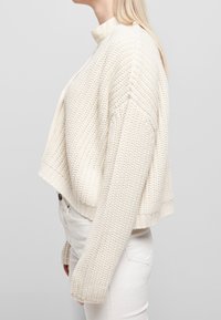Femme portant un cardigan court en grosse maille crème par-dessus un haut blanc et un pantalon blanc, vue de profil sur un fond uni.