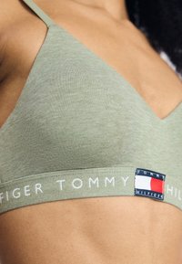 Olijfgroen bralette met een zachte, gladde textuur, V-hals ontwerp en een logoband met rode en blauwe accenten.