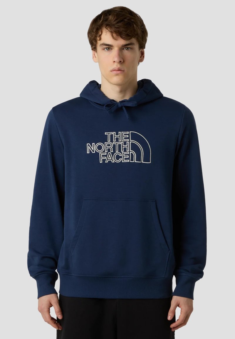 Joven con sudadera con capucha azul marino con el logo de "The North Face", de pie y mirando al frente frente a un fondo claro y liso.