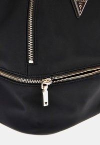 Zaino in nylon nero con zip in metallo dorato, una toppa con logo triangolare e tessuto liscio e testurizzato dal design elegante.