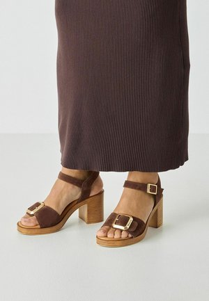 Sandales à talons en suède marron avec un bout carré, une bride de cheville ajustable et une boucle dorée. Talons carrés en bois qui ajoutent texture et hauteur.