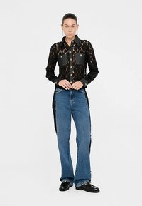 Crna čipkasta košulja sa sivim džepovima i gumbima, uparena s plavim širokim trapericama s poderanim rubom. Crne cipele upotpunjuju izgled.