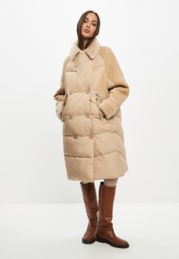 Beige quilted frakke med en skindkrave, kabelstrikkede ærmer og knaplukninger. Brune knæhøje støvler fuldender outfittet.