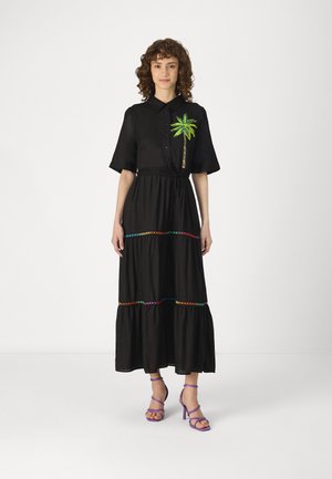 Schwarzes Maxikleid mit kurzen Ärmeln, verziert mit einer Palmenstickerei und buntem horizontalem Stich. Getragen mit lila Riemchenschuhen.