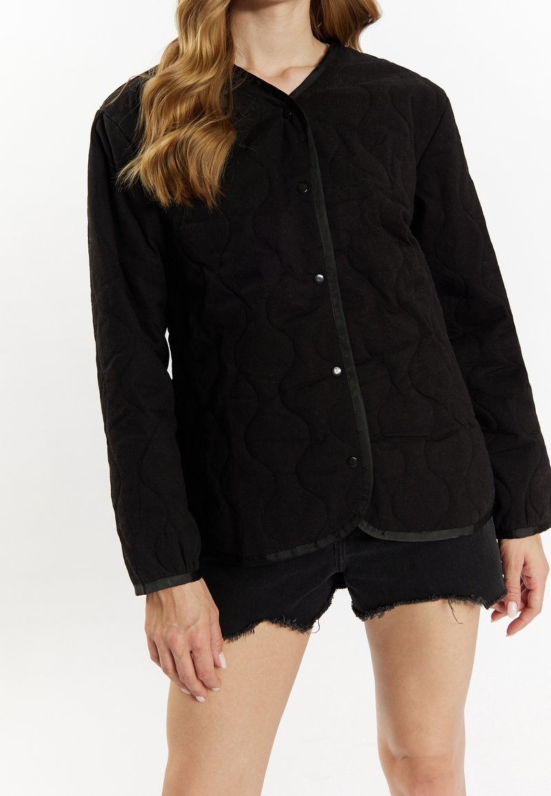 Femme aux cheveux ondulés portant une veste noire matelassée boutonnée et un short en jean noir effiloché sur un fond uni.