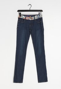 Jean slim bleu foncé avec ceinture à motif floral suspendu à un cintre noir sur fond blanc.