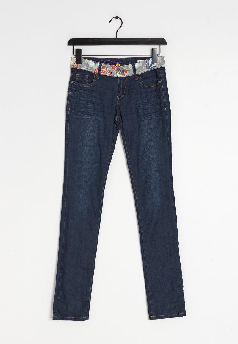 Jean slim bleu foncé avec ceinture à motif floral suspendu à un cintre noir sur fond blanc.