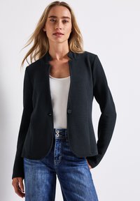 Svart texturerad blazer med rund krage, enkel knäppning och långa ärmar, matchad med en vit topp och blå jeans.