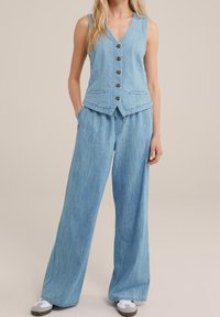 Ljusblå jeansväst med V-ringning och sex knappar, kombinerad med vida byxor. Båda plaggen har en lätt strukturerad yta.