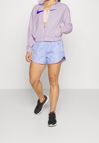 Chaqueta ligera de color púrpura con cremallera y logo de Nike en azul, combinada con pantalones cortos de color azul claro con ribetes blancos y zapatillas deportivas negras con acentos púrpuras.