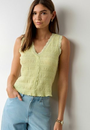 Mladá žena s hnedými vlasmi, oblečená v bezrukávovom textúrovanom svetložltom topánku na gombíky a svetlomodré džínsy, stojaca pri bielom múre.