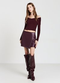 Burgunderfarbener, gerippter Off-Shoulder-Crop-Top kombiniert mit einem figurbetonten Leder-Minirock mit Druckknöpfen, kniehohen glänzenden Stiefeln und einer eleganten Silhouette.