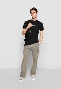 Black cotton t-shirt with printed graphic, tan cargo pants with side pockets, and white sneakers. Accessories include a black crossbody bag.