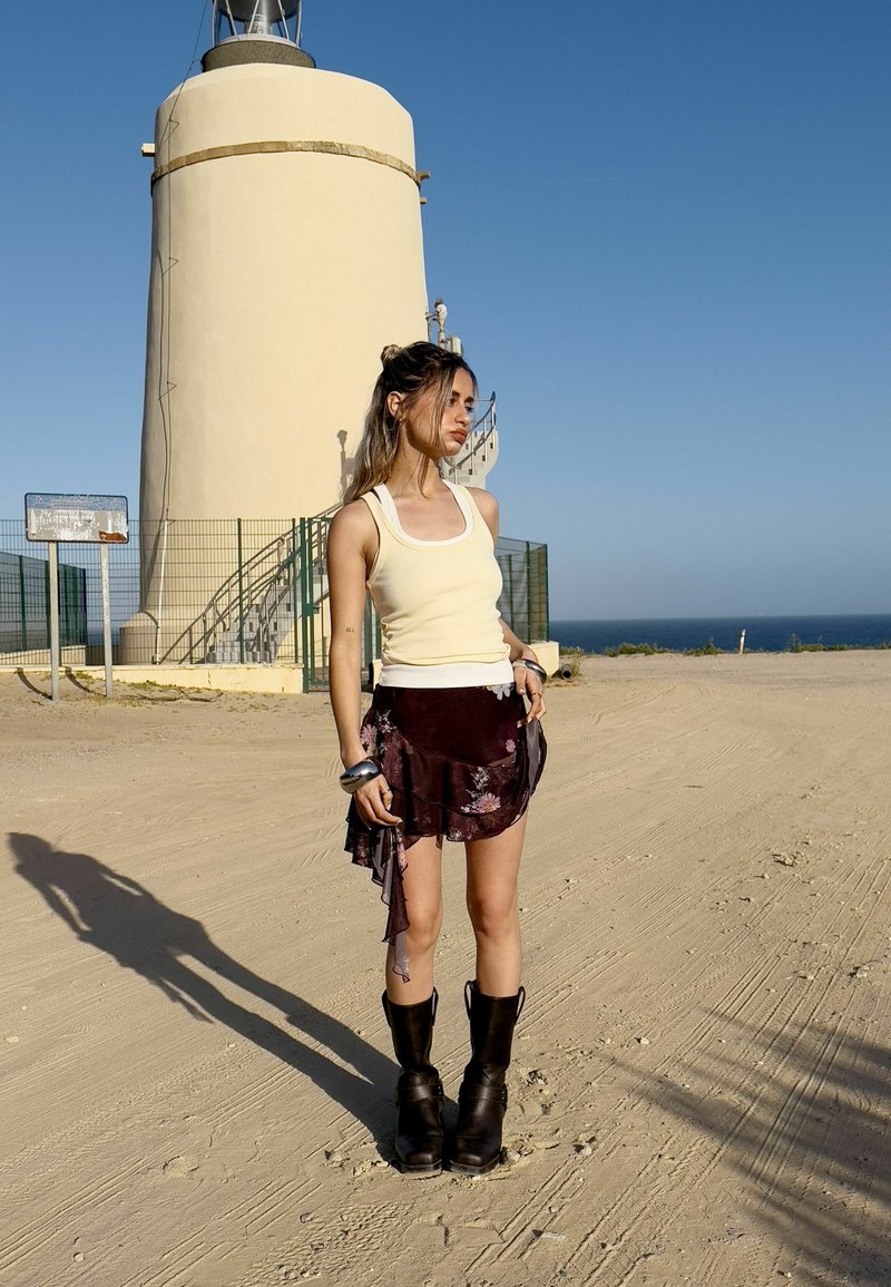 Débardeur jaune, jupe florale bordeaux superposée, bottes noires jusqu'aux genoux. L'arrière-plan présente un phare de couleur crème sous un ciel bleu clair.