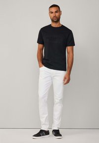 Schwarzes T-Shirt mit kurzen Ärmeln und geprägtem "HACKETT SPORT"-Logo, kombiniert mit weißen Slim-Fit-Jeans und schwarzen Sneakers vor einem neutralen Hintergrund.