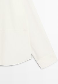 Manche de chemise blanche à manches longues avec un détail de poche et une fente latérale à l'ourlet sur un fond uni.