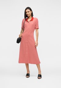 Γυναίκα που φοράει midi φόρεμα με ρίγες κόκκινου και άσπρου, μαύρα σανδάλια slide και μαύρη τσάντα χιαστί, ποζάροντας μπροστά σε λευκό φόντο.