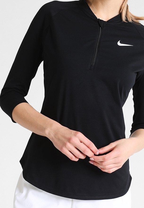 Kobieta ubrana w czarną sportową bluzę z długim rękawem i białym logo Nike, z półzamkiem przy kołnierzu, oraz białe spodnie, z rękami złożonymi na wysokości talii.