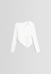 Vit långärmad kort blus med en asymmetrisk knappsydd framsida och en spetsig fåll på en ljusgrå bakgrund.