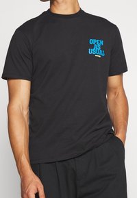 Svart bomulls t-shirt med blå och gula texter "ÖPPET SOM VANLIGT" på framsidan. Rund halsringning och korta ärmar. Slät textur.