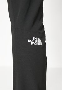Lähiprofiili mustista housuista, joissa on valkoinen "The North Face" -logo reiden kohdalla tasaisella taustalla.