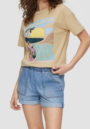 Femme portant un t-shirt graphique beige avec un motif coucher de soleil et océan et un short en denim bleu clair roulé sur un fond uni.