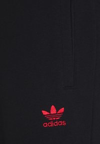 Černá látka s červeným vyšívaným logem Adidas trefoil blízko vertikálního švu nebo zipu.